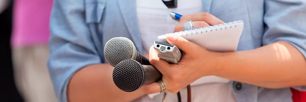 jornalista anotando em caderneta e segurando microfones e gravador
